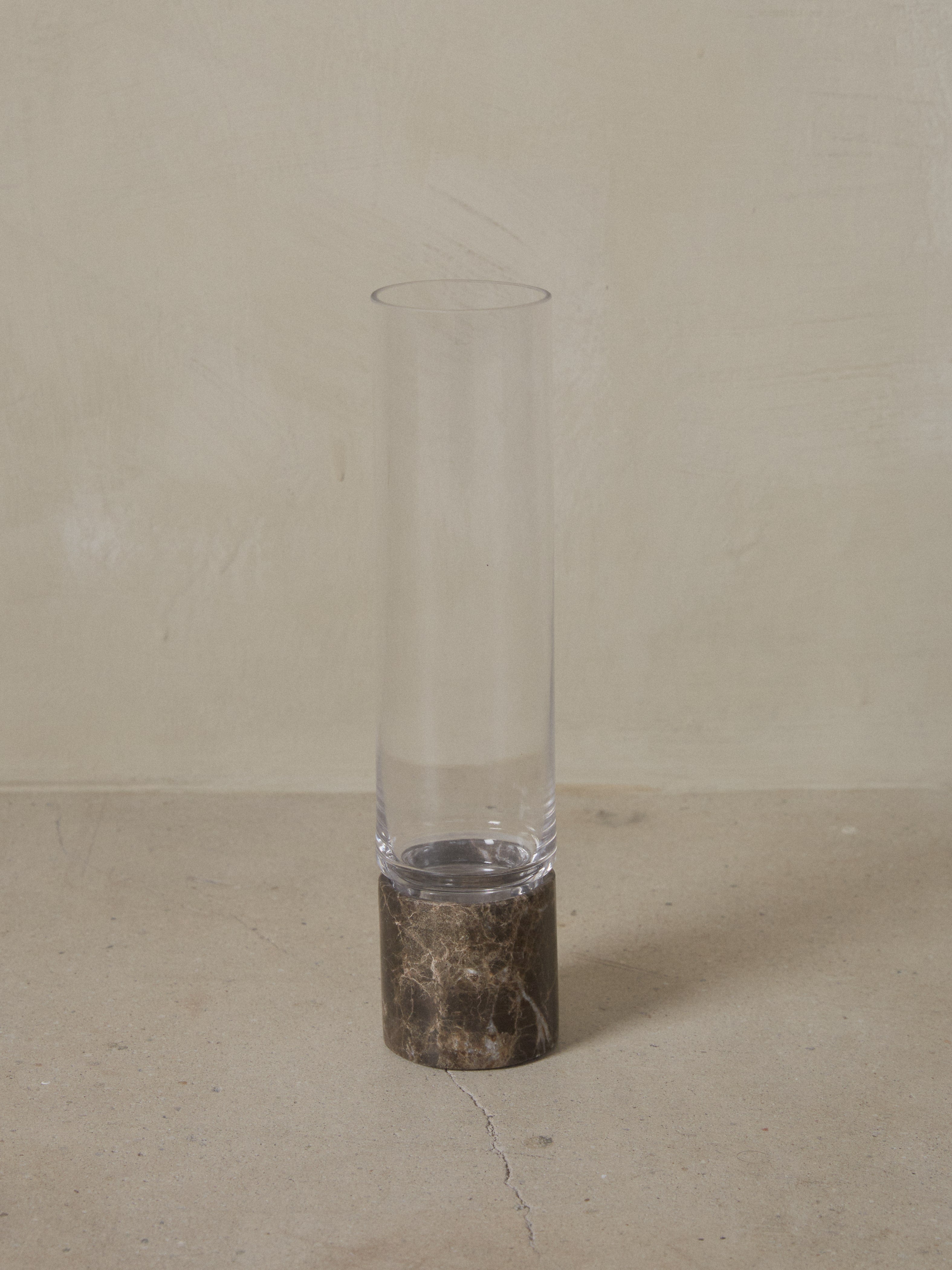 A minimal handblown cylindrical glass vase with a lean silhouette rests atop an elegant, solid Dark Emperador marble base. 