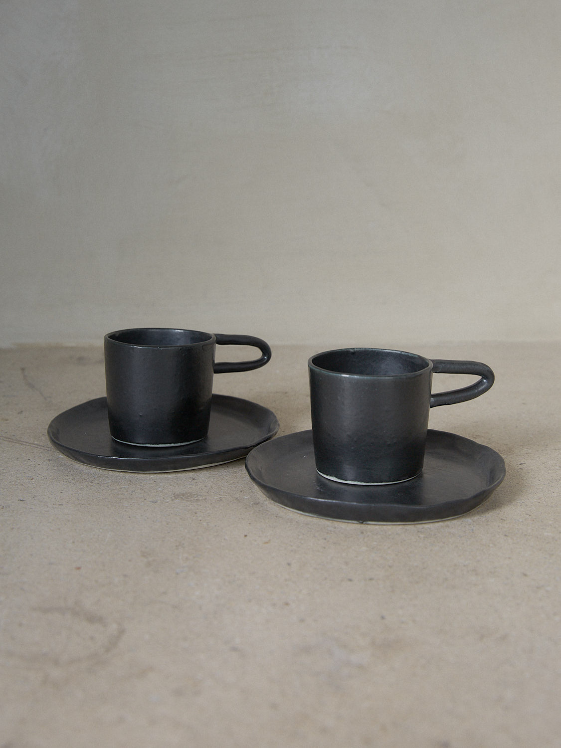 Black Raw Espresso Cup Set. A set of two moody, handmade espresso cups with linear extended handles and matching saucers in matte black stoneware.