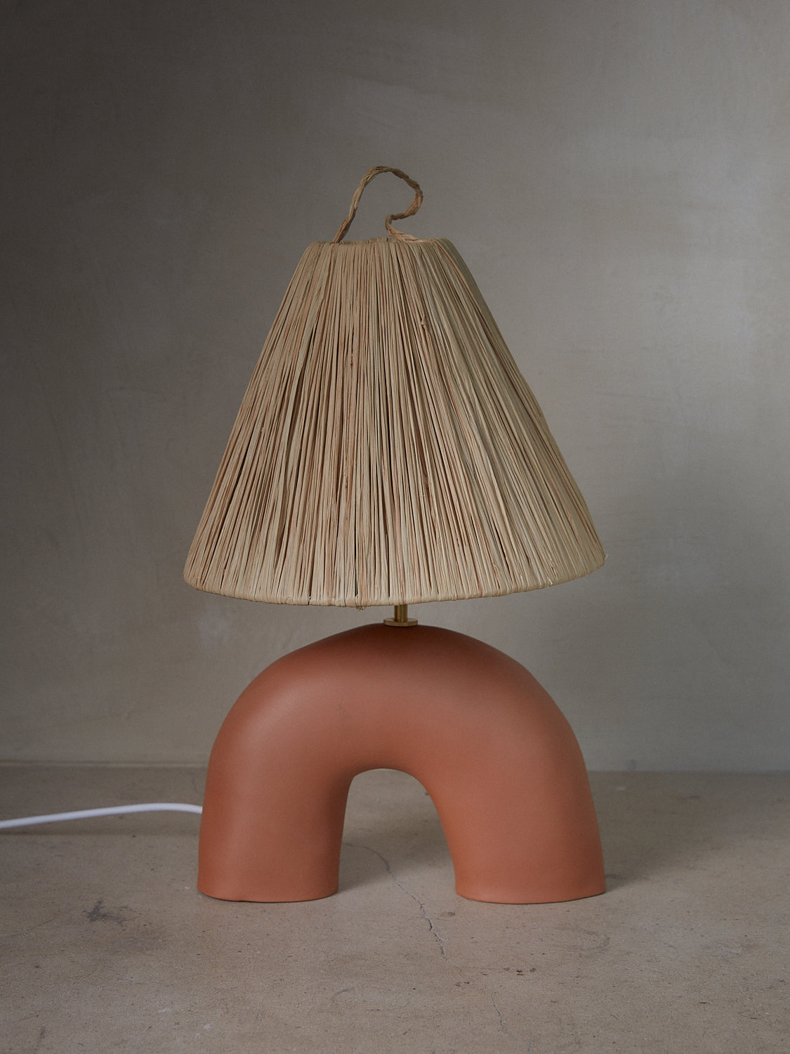 Terracotta Volta Lamp. Playful, handmade terracotta lamp with a burnished finish, brass hardware, cotton wiring and charming raffia shade